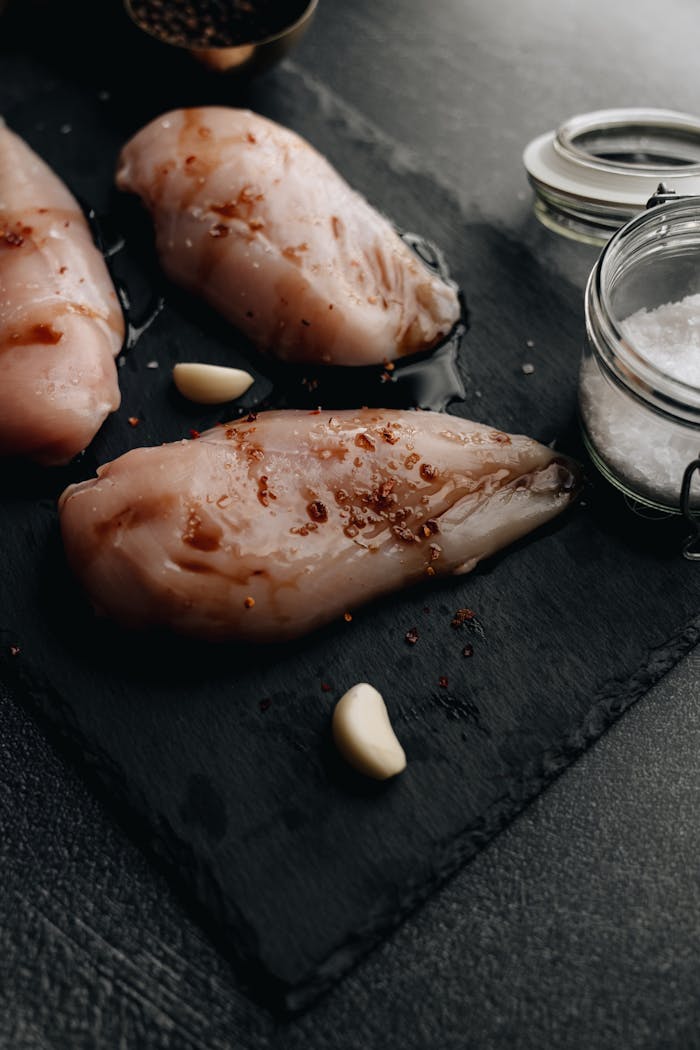 Close-up of raw marinated chicken breasts with garlic and salt on a dark slate board.
